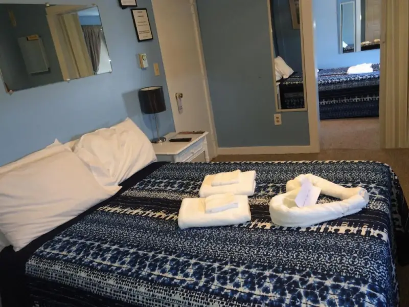A neatly made bed with white pillows and a blue patterned comforter, with folded towels on top and a mirror on the wall.