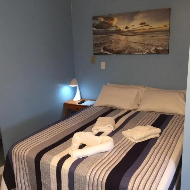 A neatly made bed with a striped comforter and towels on top, a nightstand with a lamp, and a painting of a beach scene on the wall.